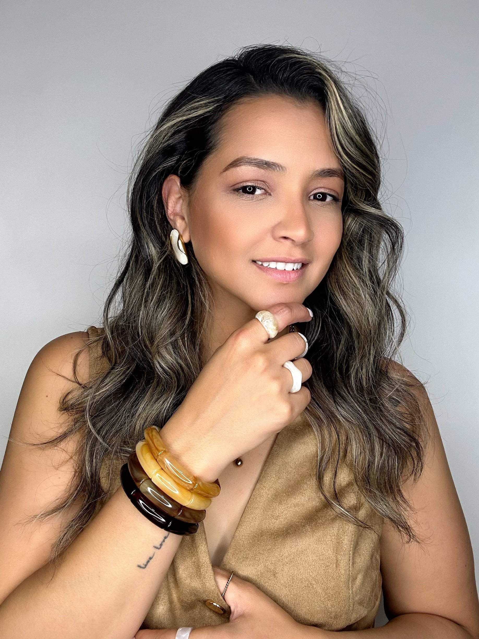 Woman with long, wavy hair wearing multiple bracelets and a ring against a neutral background